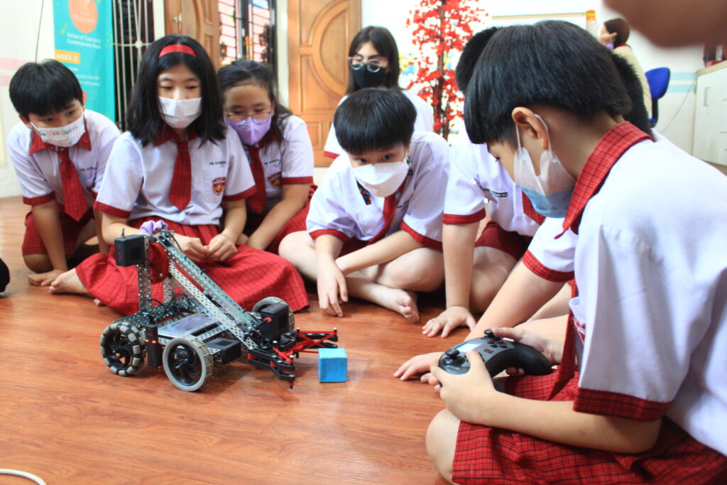 Anak sekolah dasar mengendalikan robot penjepit menggunakan remote. Contoh edukasi dan inovasi teknologi robotik untuk anak usia dini.